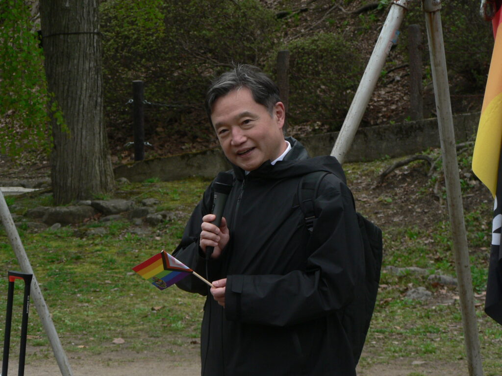 Morioka City Mayor Uchidate Shigeru speaks into a microphone while holding a small Progress Pride flag