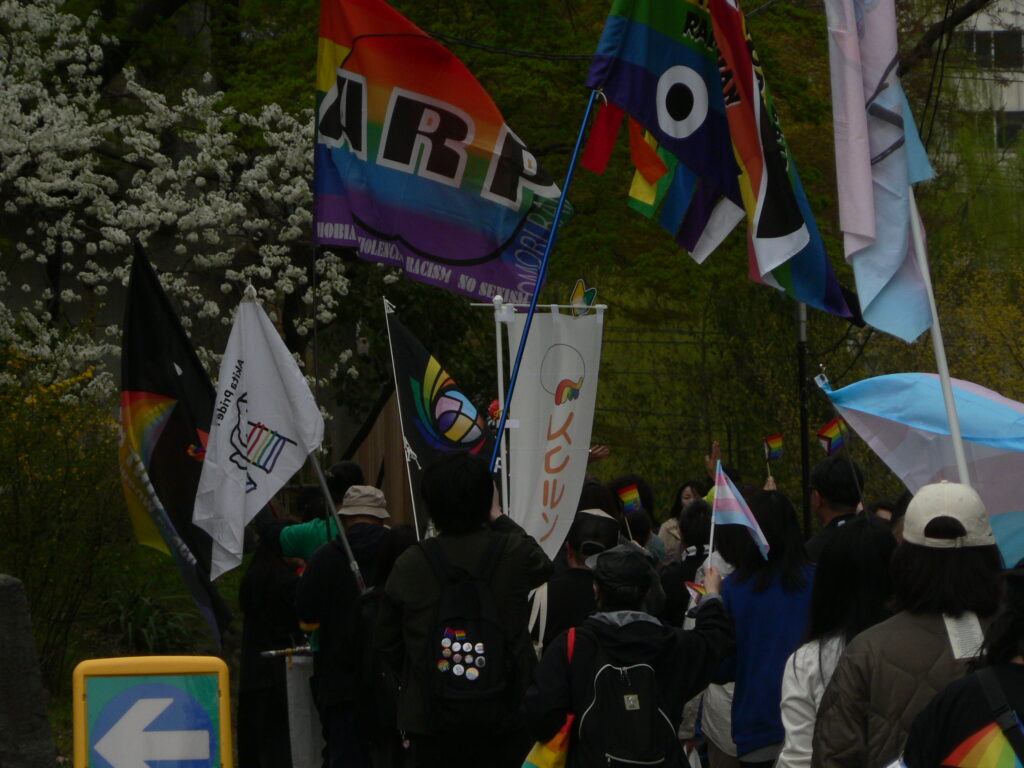 Pride marchers wave rainbow, trans, and organization flags as they pass blooming cherry trees in Morioka