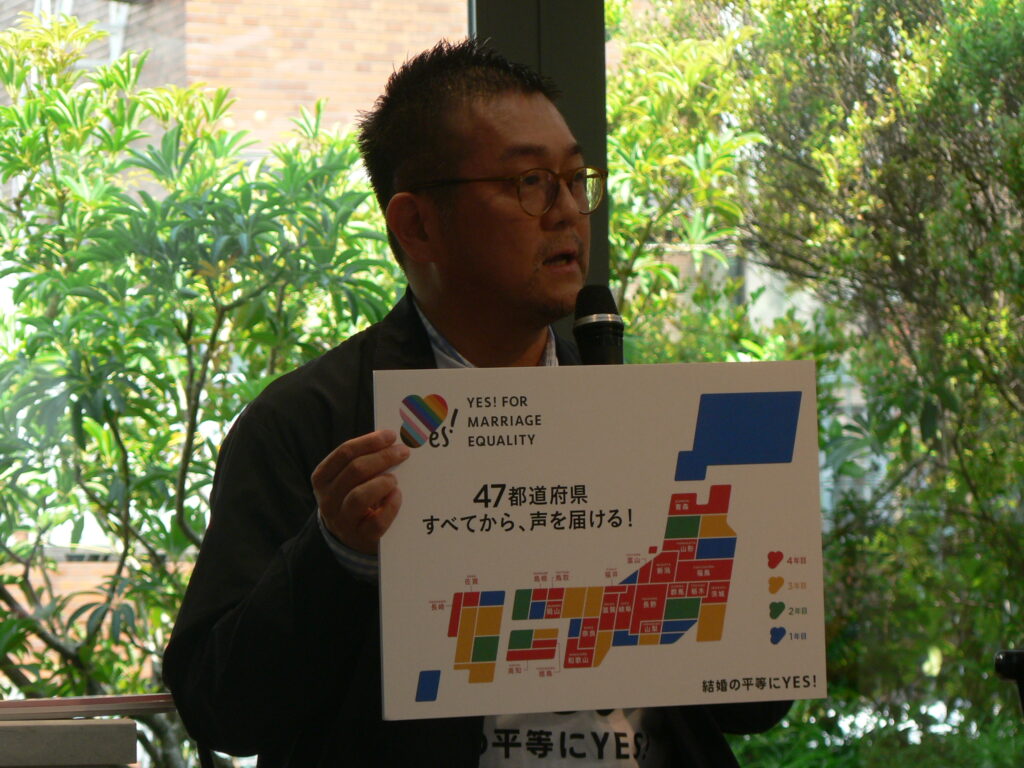 Matsunaka Gon speaks into a microphone holding a placard with a Japan map and the campaign's slogan about reaching all 47 prefectures.