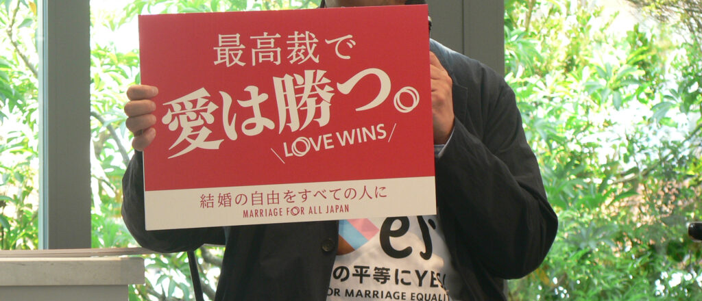 Matsunaka Gon holds a red placard reading 'Love Wins' in Japanese and English, with the Marriage for All Japan slogan beneath.