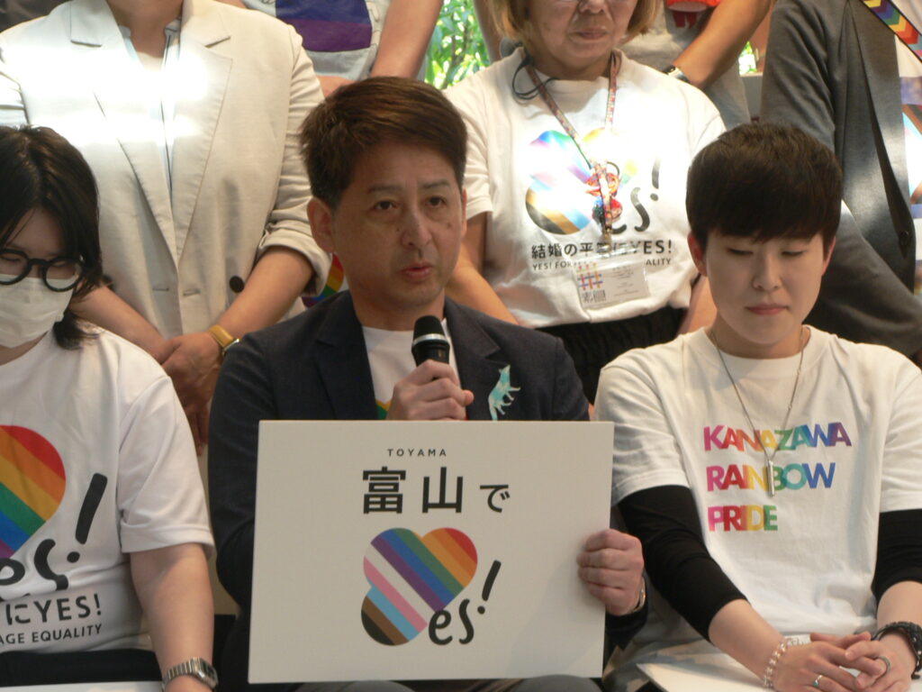 A man speaks into a microphone holding a 'Toyama, Yes!' placard, seated among supporters in rainbow marriage-equality shirts.