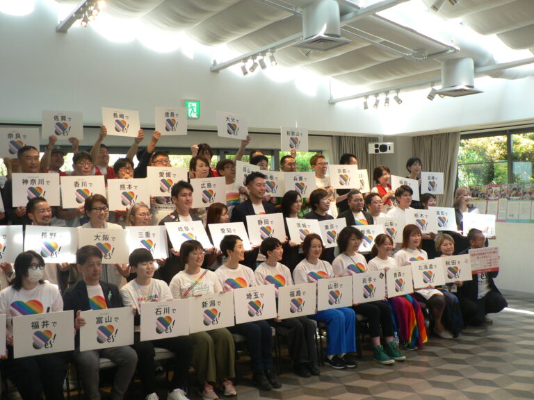 About fifty people hold white placards naming Japanese prefectures, each with a rainbow 'YES!' marriage-equality logo.