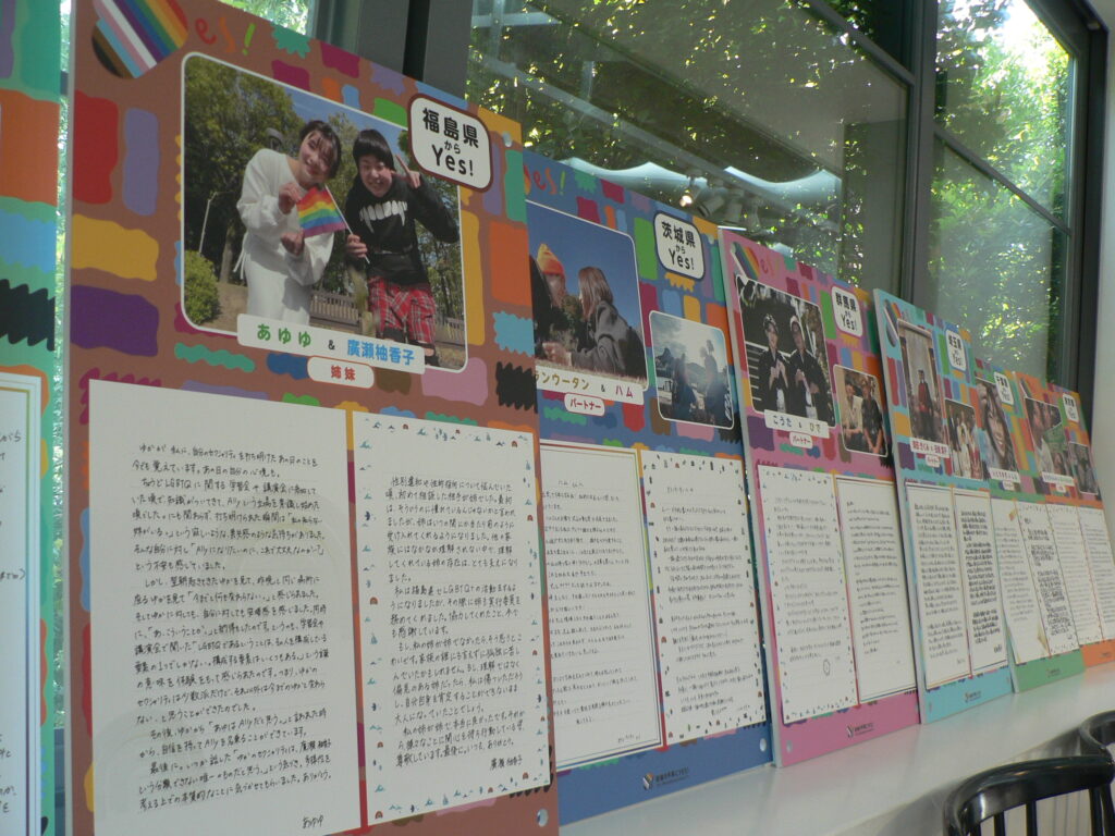 Colorful exhibition panels along a window, each with a couple's photo, a prefecture 'Yes!' label and handwritten letters.