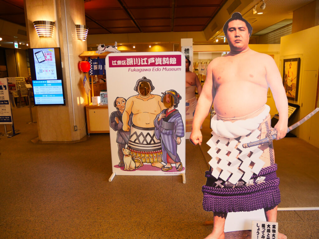Life-size sumo wrestler cutout in a ceremonial apron beside a Fukagawa Edo Museum sign