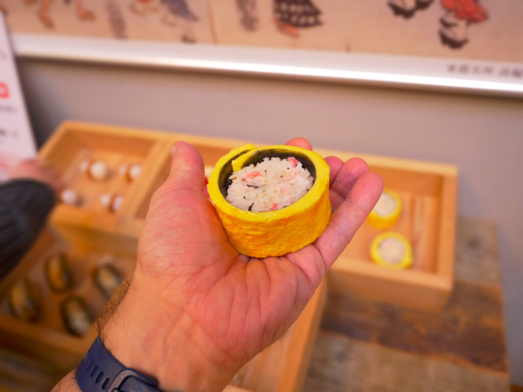 A hand holds a plastic replica of oversized Edo-era sushi above museum display trays of food models