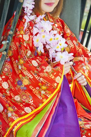 Woman in a vibrant red jūnihitoe holding cherry blossoms, with colorful layered robes visible beneath