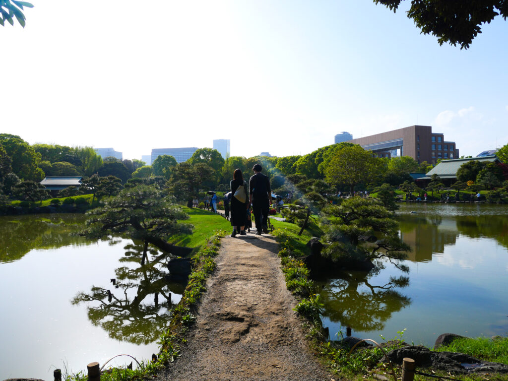 Two visitors walk a stone causeway across Kiyosumi Garden's pond, framed by low pines with Tokyo buildings beyond