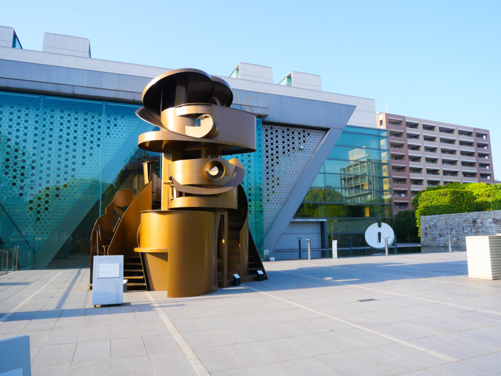 A large bronze spiral sculpture in the plaza outside the angular glass facade of MOT Tokyo