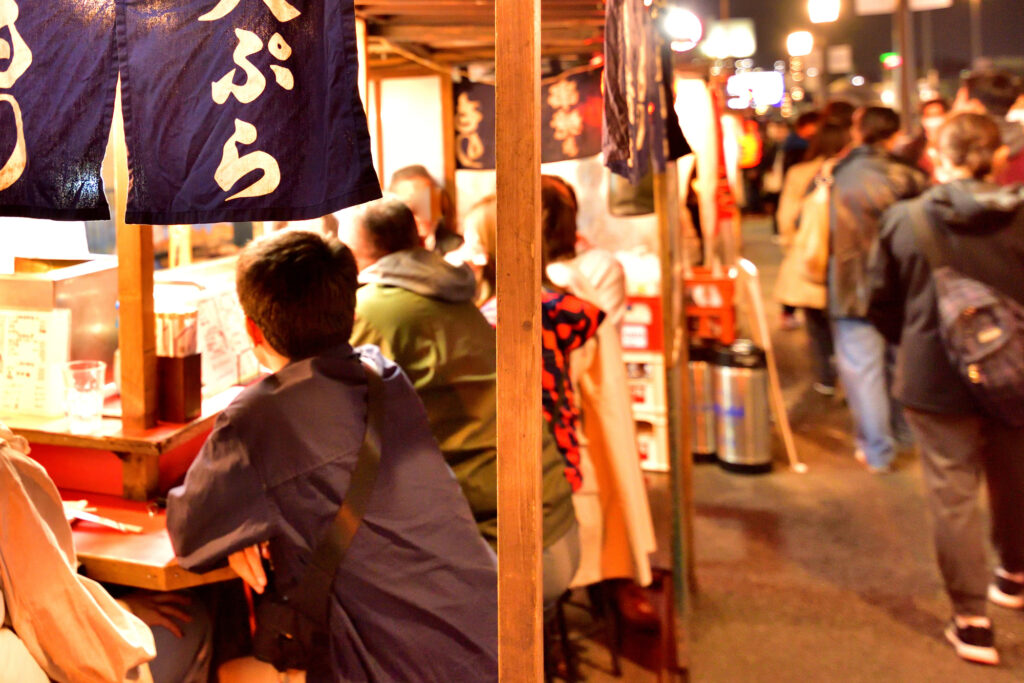 Diners eat at a Fukuoka yatai food stall at night, hung with a noren curtain reading tempura.