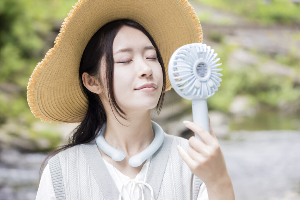 Woman wearing a straw hat and cooling neck ring while holding a handheld electric fan.