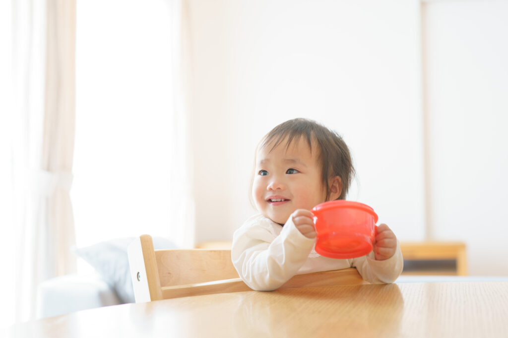 Toddler in a high chair smiling while holding a red cup at a dining table