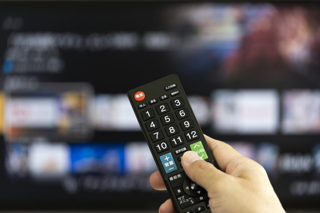 Hand holding a Japanese TV remote in front of a blurred television screen