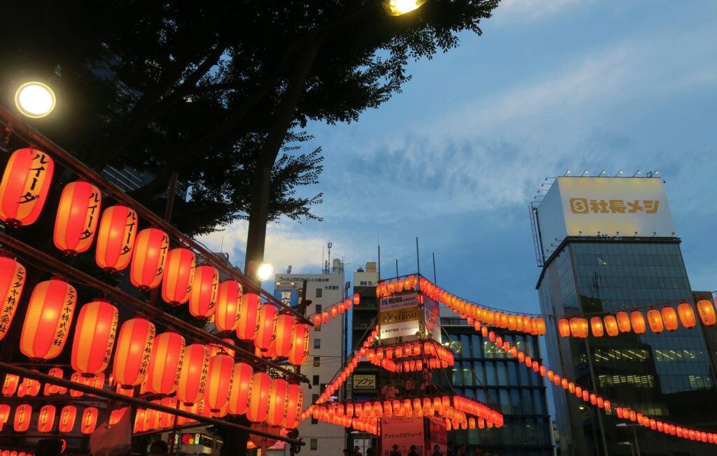 Rows of glowing red paper lanterns strung across an urban Bon Odori venue at dusk