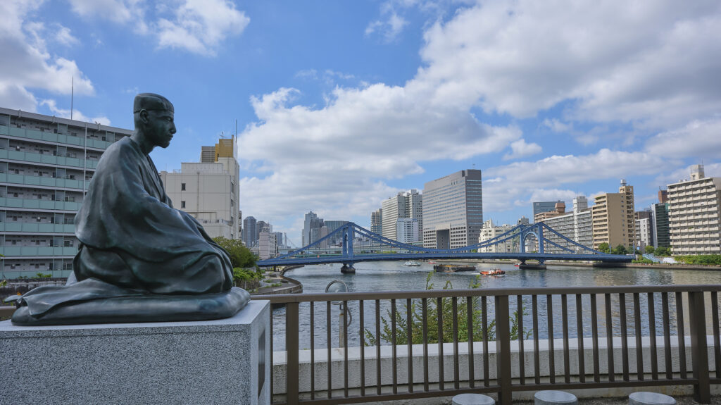 Bronze seated statue of the poet Bashō overlooking the Sumida River and the blue Kiyosu Bridge