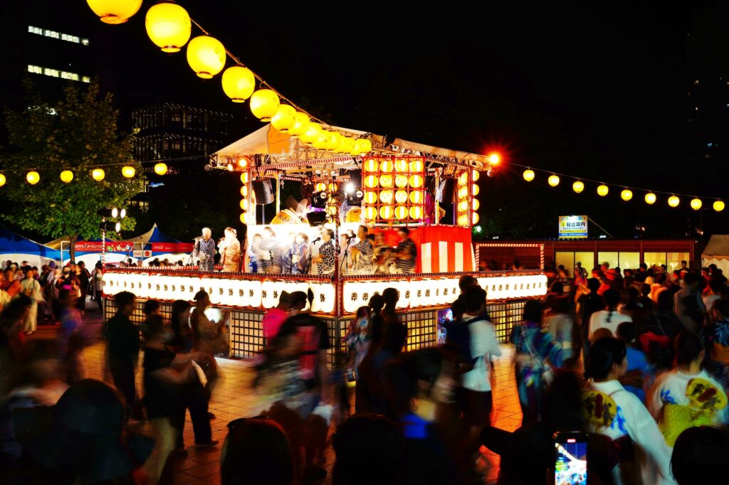 Crowds dancing around an illuminated yagura stage at a nighttime Bon Odori festival
