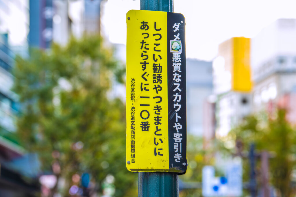 Yellow Japanese street sign warning against aggressive scouts and touts, urging passersby to call 110 if harassed