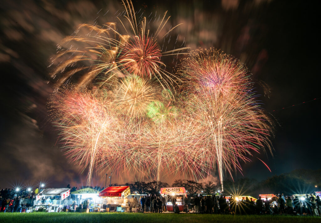 Fireworks light up the sky above yatai food stalls as spectators watch from a grassy field at Tsuchiura