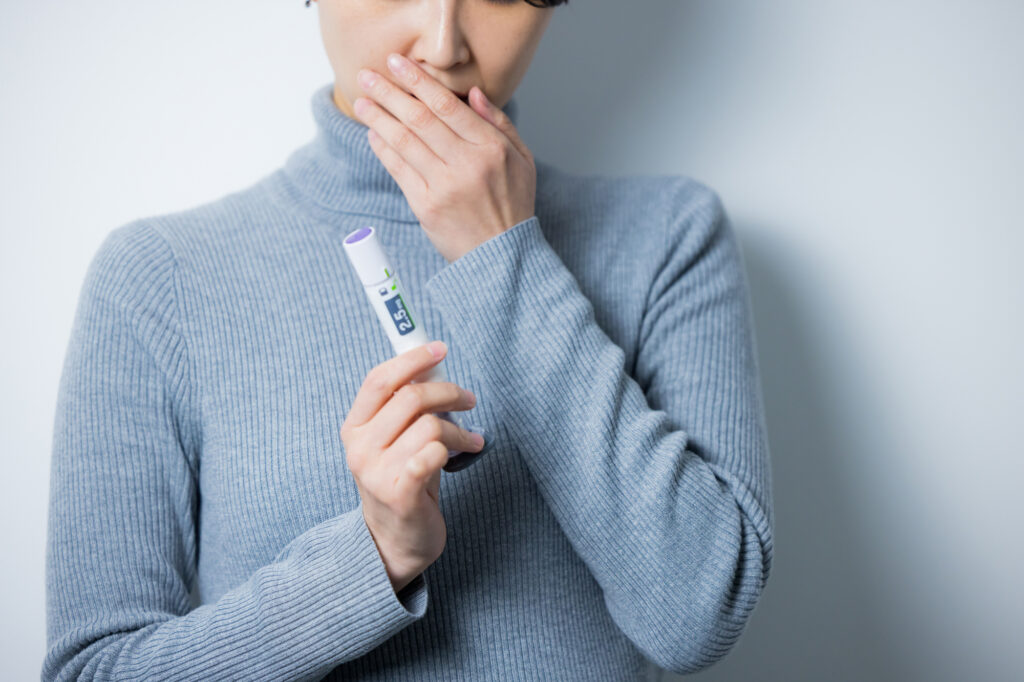 A woman in a gray turtleneck holds a GLP-1 injection pen near her face, looking apprehensive.