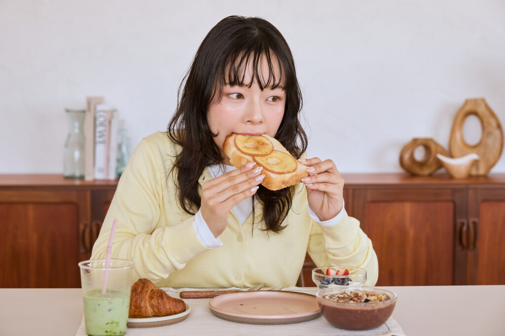 Woman biting into a thick slice of toast at a breakfast table with a croissant, smoothie, and granola