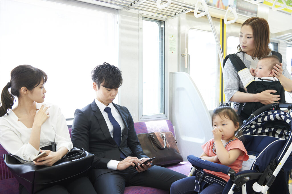 Mother stands on a crowded Japanese train holding a baby in a carrier with a toddler in a stroller beside seated commuters