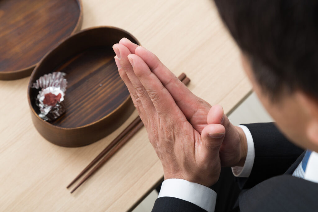 Man in a suit pressing his palms together over an open wooden bento box with an umeboshi, chopsticks beside it