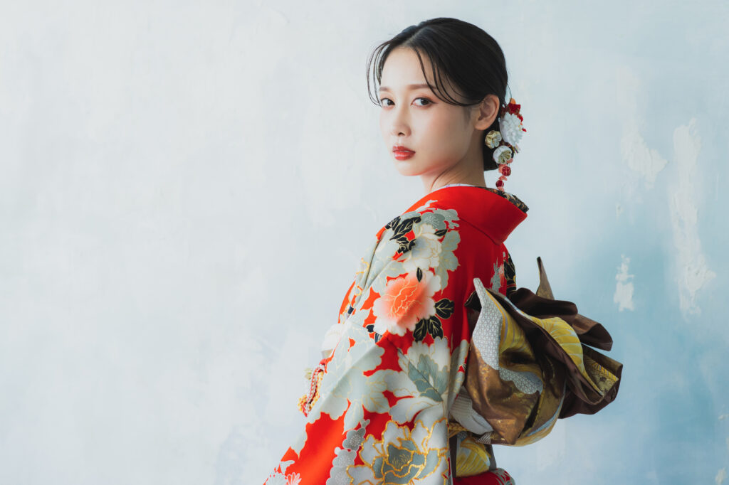 Young woman in a red furisode with bold peony motifs and a gold obi, glancing over her shoulder
