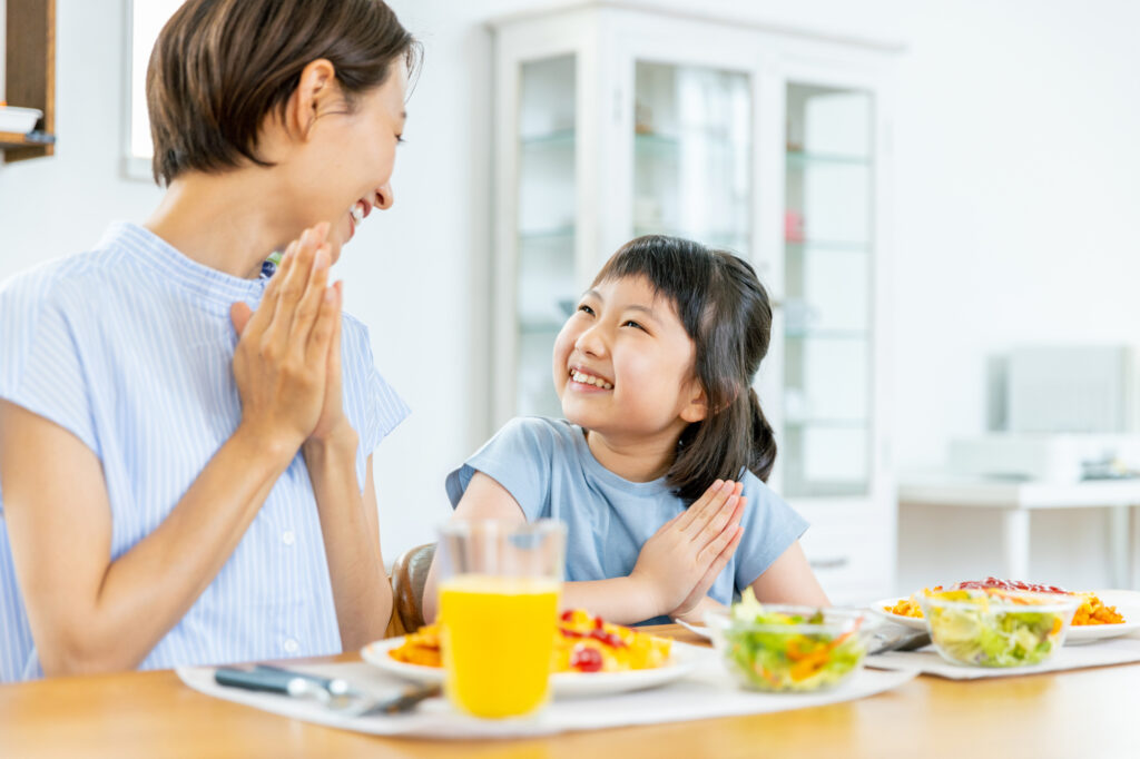 Mother and young girl press their palms together to say itadakimasu before a home meal, smiling at each other