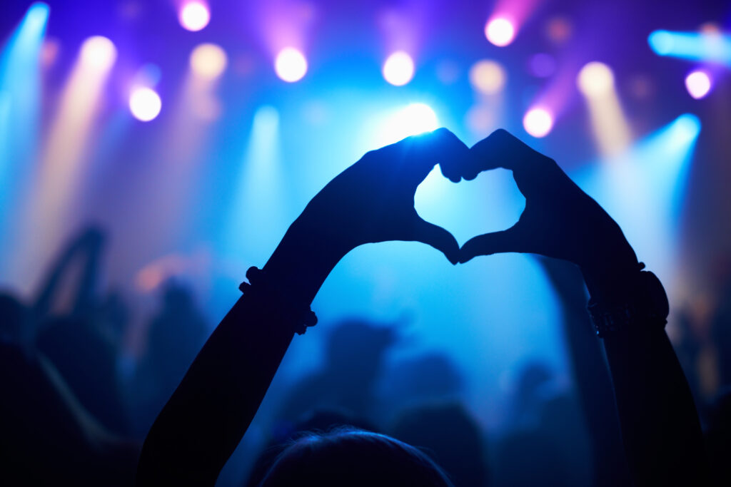 Silhouette of a fan's hands forming a heart shape against blue and purple concert stage lights