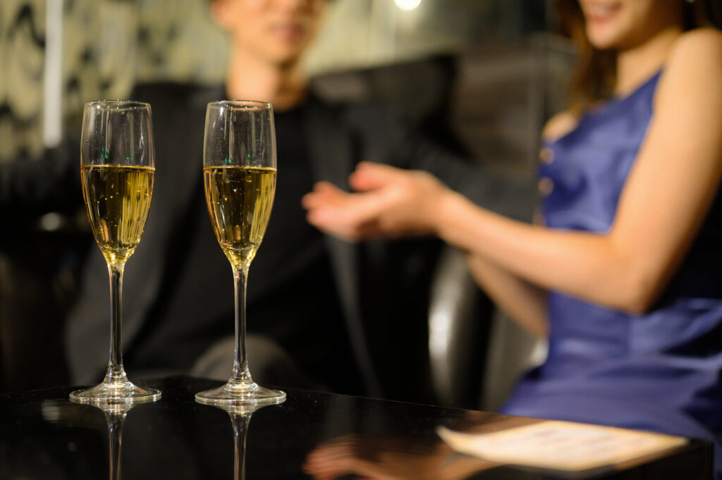 Two champagne flutes on a bar table with a couple talking in the background