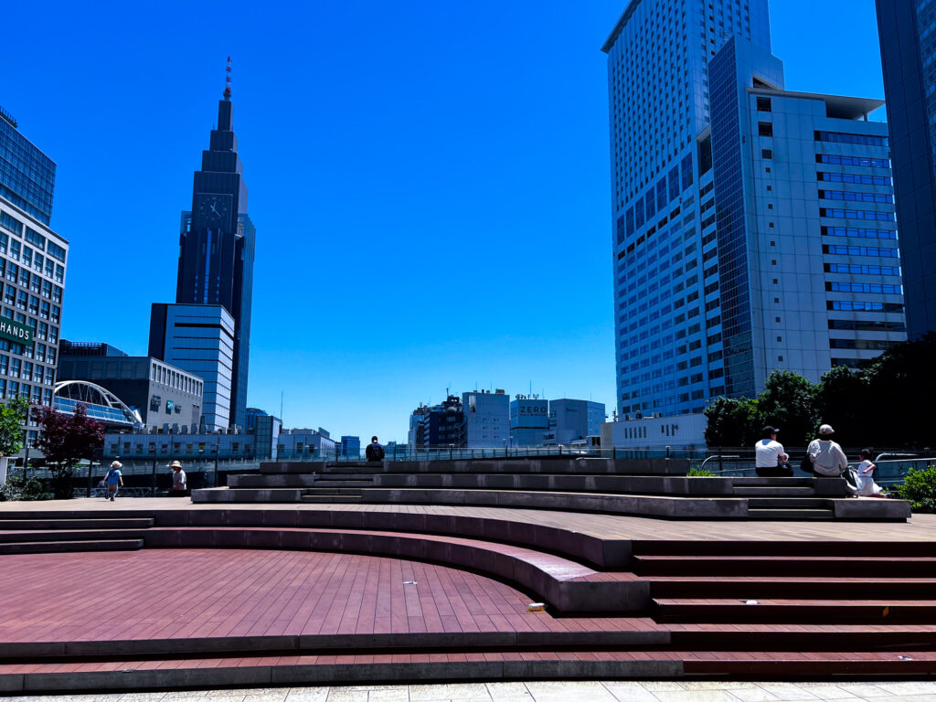Outdoor tiered seating area near Shinjuku Station on a clear day, with the NTT Docomo Yoyogi Building in the background