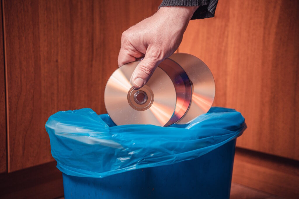 A hand dropping compact discs into a blue plastic-lined trash bin