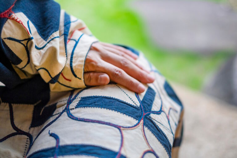 Person in a traditional Ainu robe with indigo and white appliqué patterns, one hand resting on the knee