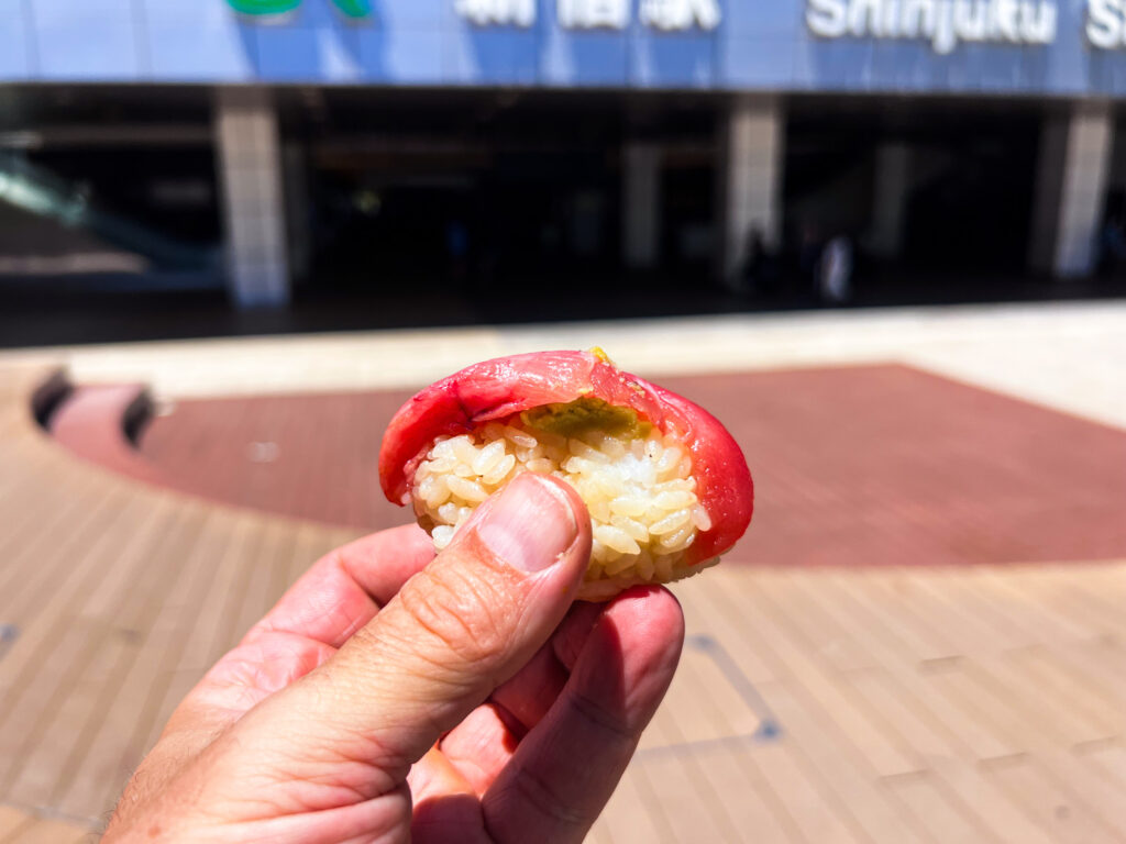 Partially bitten tuna Edomae sushi showing rice and wasabi inside, held up outside Shinjuku Station