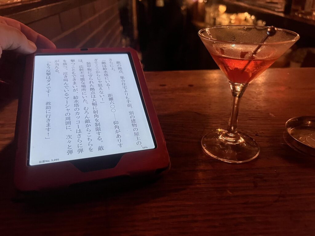 A red cocktail in a stemmed glass on Bar Lupin's wooden counter, beside an e-reader showing Japanese text