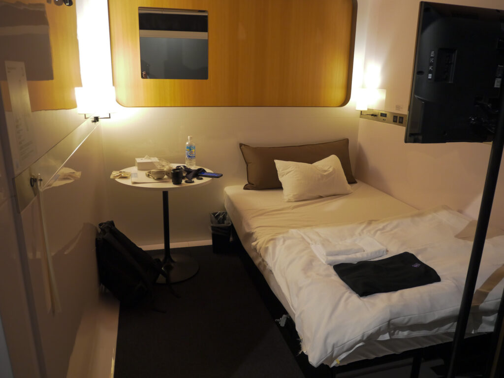 A roomier First Class cabin with a single bed, a round side table, a wall-mounted TV, and floor space for luggage