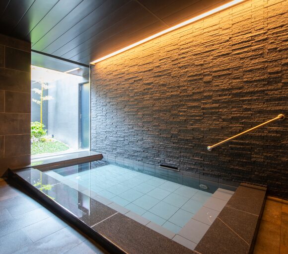 A small indoor artificial hot-spring bath with stone-tiled walls, a brass handrail, and a window onto a garden