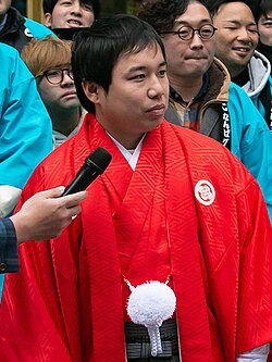 Comedian Seiya in a red formal haori speaks into a handheld microphone at an outdoor event
