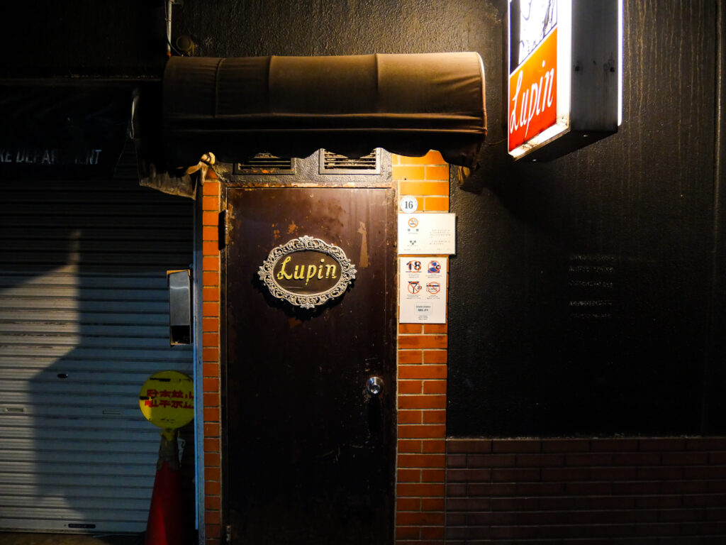 Close-up of Bar Lupin's weathered wooden door with an oval Lupin nameplate and brick surround