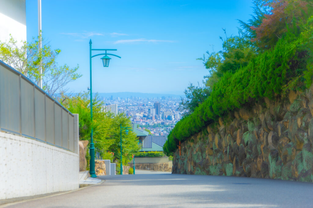 A quiet hillside street in Ashiya descends past stone retaining walls toward a sweeping city and bay view.