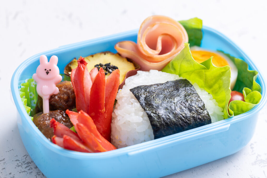 Child's blue bento with a nori-wrapped onigiri, flower-cut sausages, a ham rosette, meatballs, egg, and a rabbit food pick