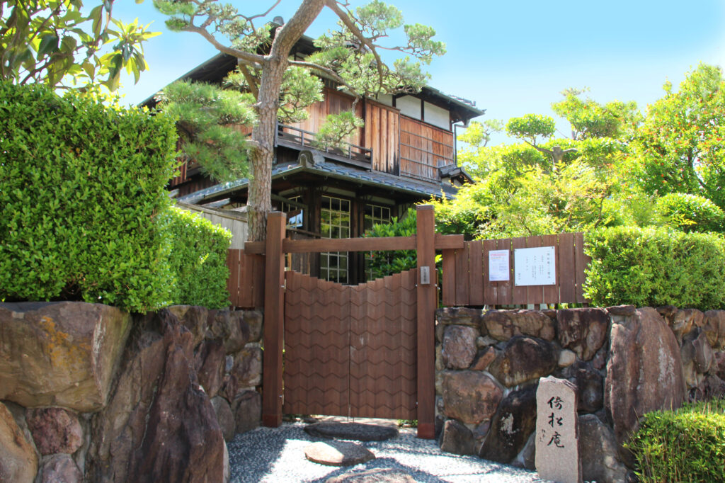 A traditional wooden Japanese house behind a wooden gate and stone wall, framed by a pine tree and hedges.