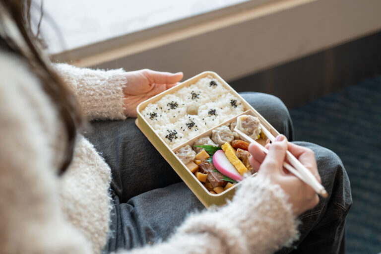 Person eating a Japanese bento of sesame-topped rice, shumai, tamagoyaki, and kamaboko with chopsticks on their lap