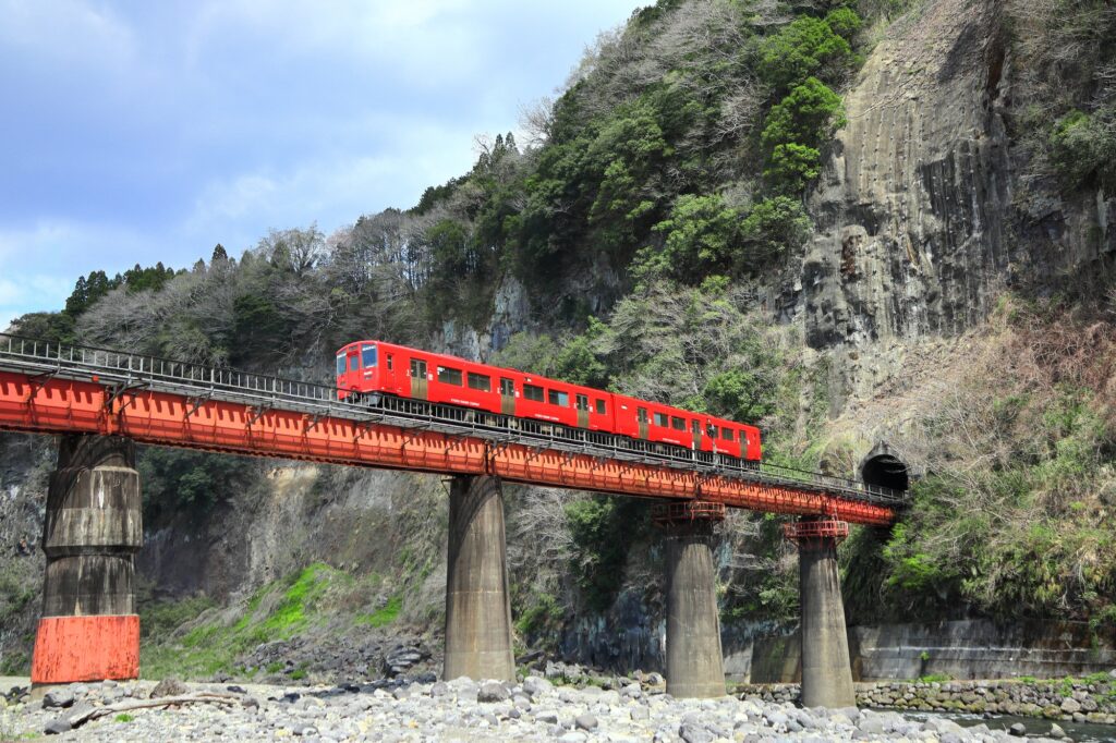 A red local train crossing a red steel railway bridge over a rocky river in a mountainous gorge in Ōita, Kyushu
