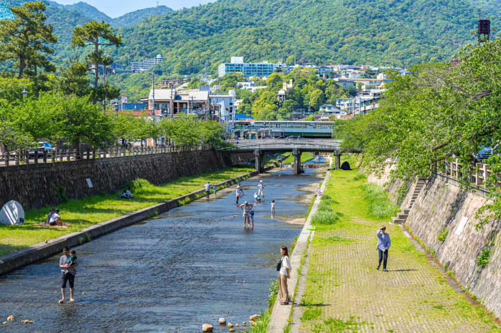 Walkers on the paved banks of the Ashiya River in summer, with the green Rokko Mountains behind the town.