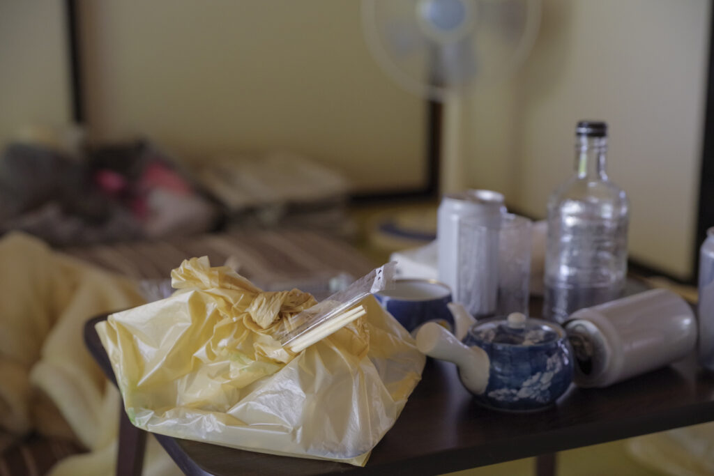 A low table cluttered with empty cans, a bottle, and leftover food beside an unmade futon