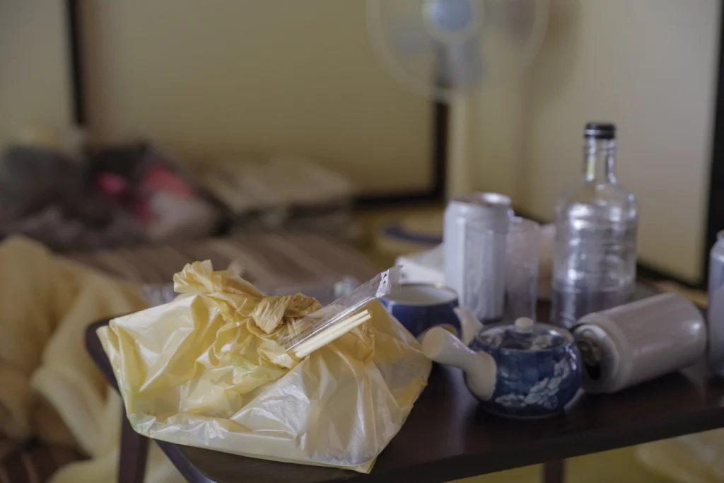 A low table cluttered with empty cans, a bottle, and leftover food beside an unmade futon