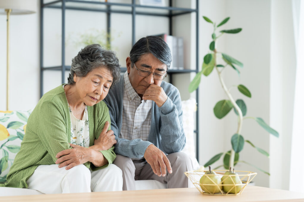 An elderly couple sit close together looking down with worried, downcast expressions