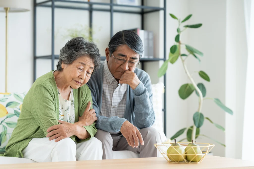 An elderly couple sit close together looking down with worried, downcast expressions