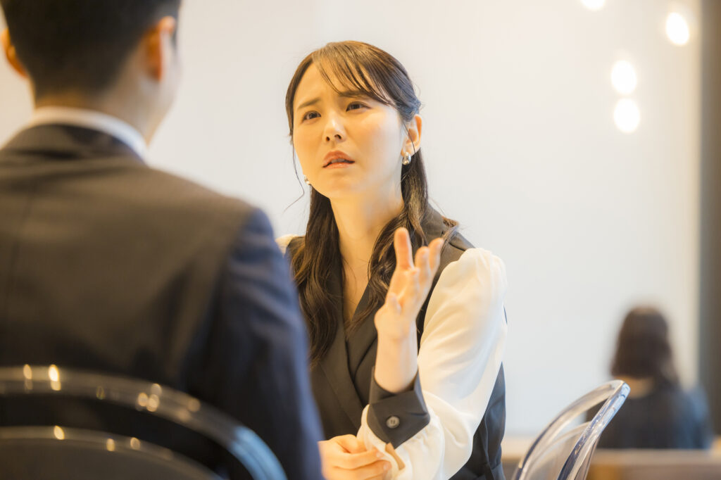 Woman gesturing with a troubled expression toward a man in a suit seen from behind across a table