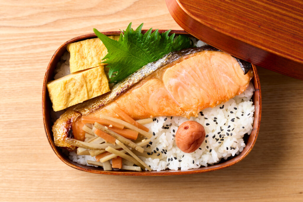 Open wooden bento box with grilled salmon over rice, an umeboshi plum, tamagoyaki, and burdock kinpira